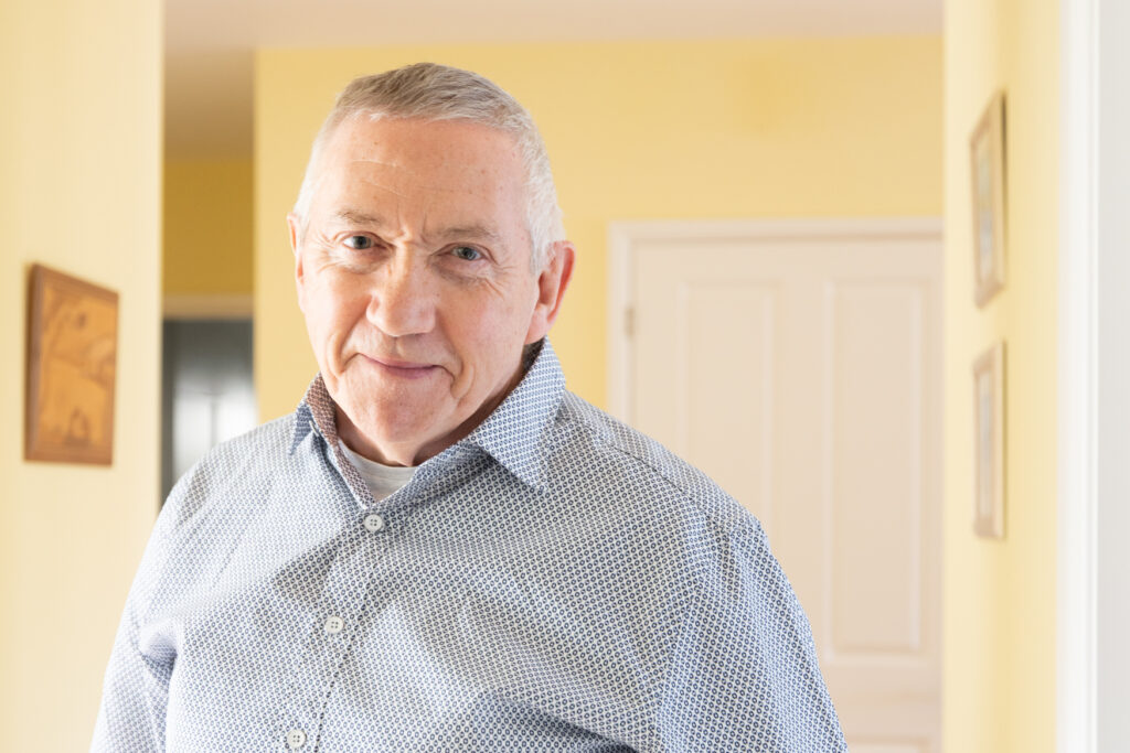 Older man in blue shirt standing indoors, yellow walls and white door behind, serious expression, composer or theatre professional possibly