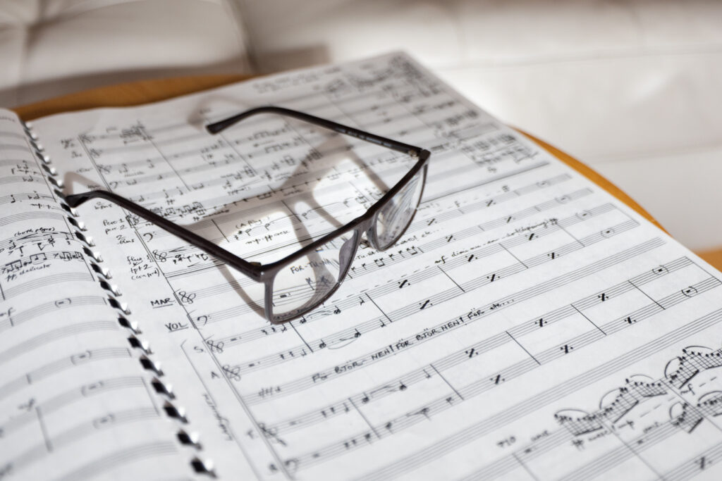 Glasses on sheet music, black frames on white pages with notes, on a wooden table in a bright room, suggesting a composer at work in a theatre or studio setting